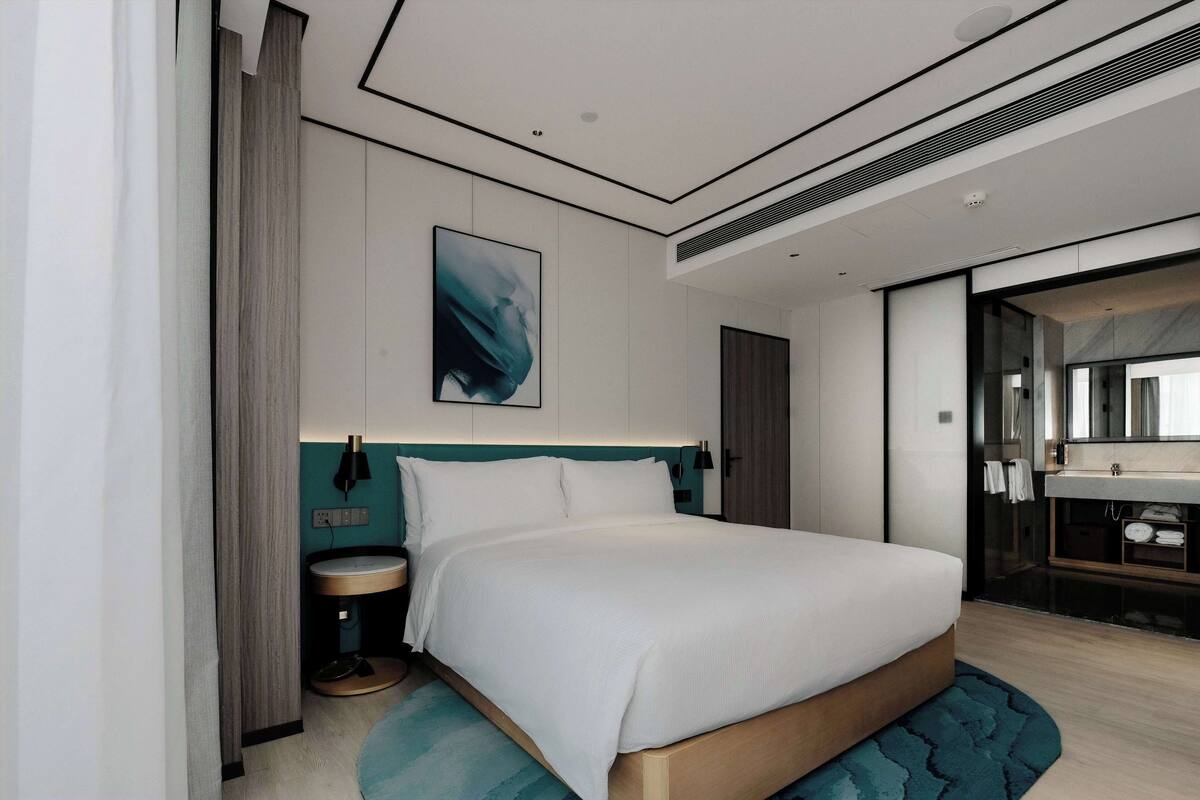 A modern hotel room with a large bed, a bedside table, a built-in vanity with a mirror, and a large window.