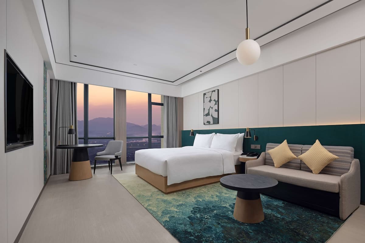 A modern hotel room with a large bed, a sofa, a dining table, and a view of the cityscape through large windows.