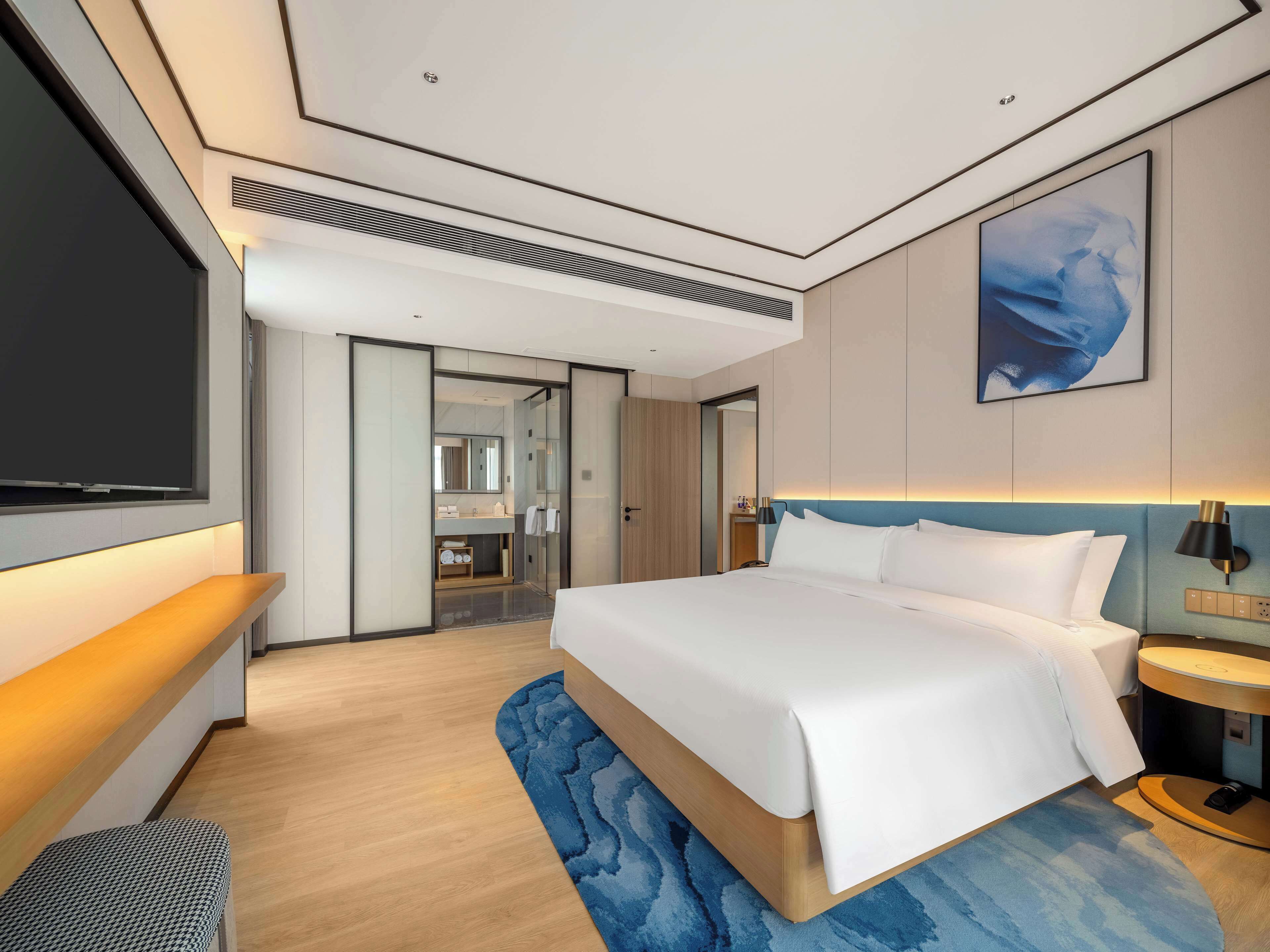 Una habitación de hotel moderna con una cama grande, un televisor, un baño con espejo y una pared azul decorativa.