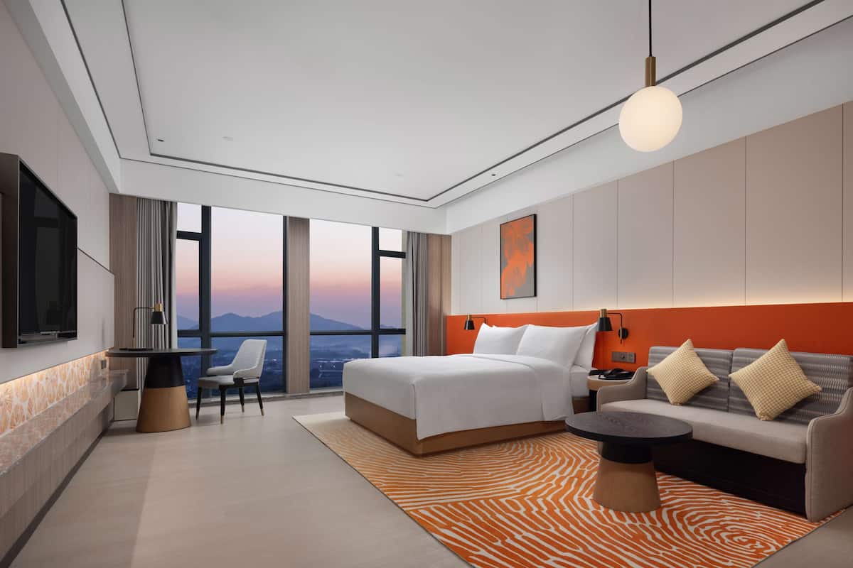 A modern hotel room with a large bed, a sofa, a desk, and a view of the mountains.
