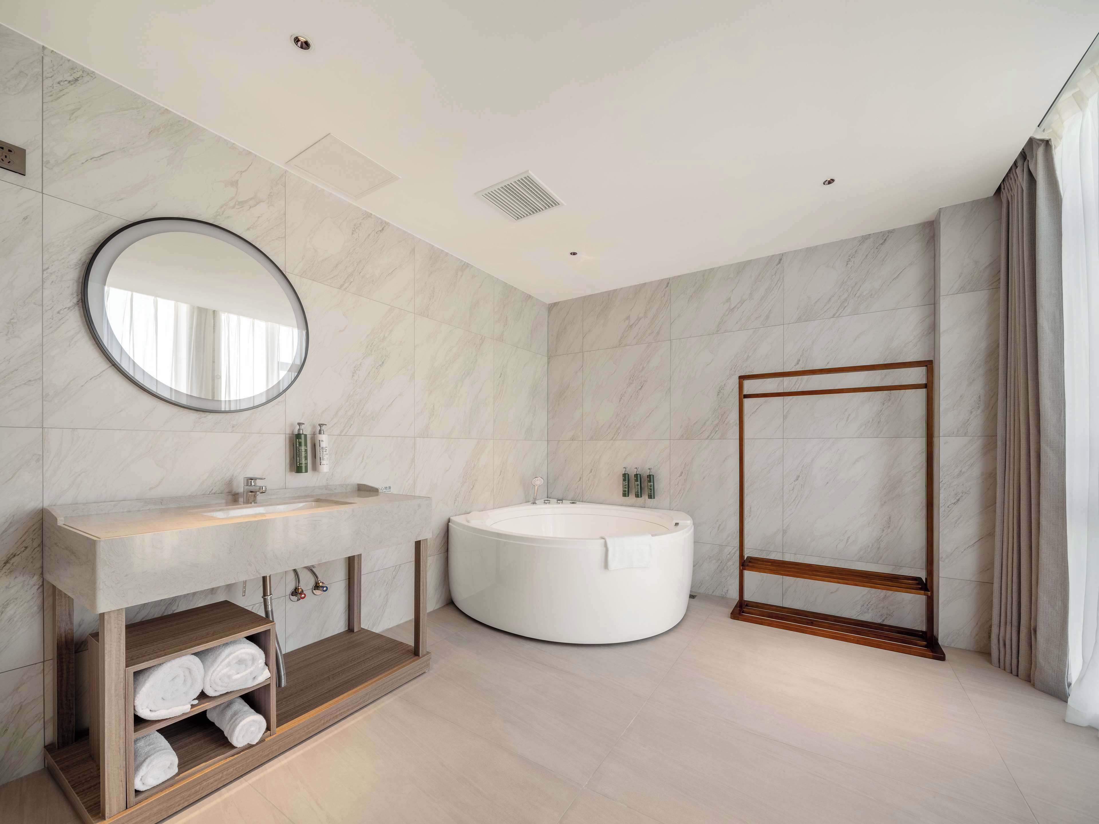 Un baño moderno con suelo de mármol, un espejo redondo, una bañera independiente y un mueble de tocador de madera con toallas.