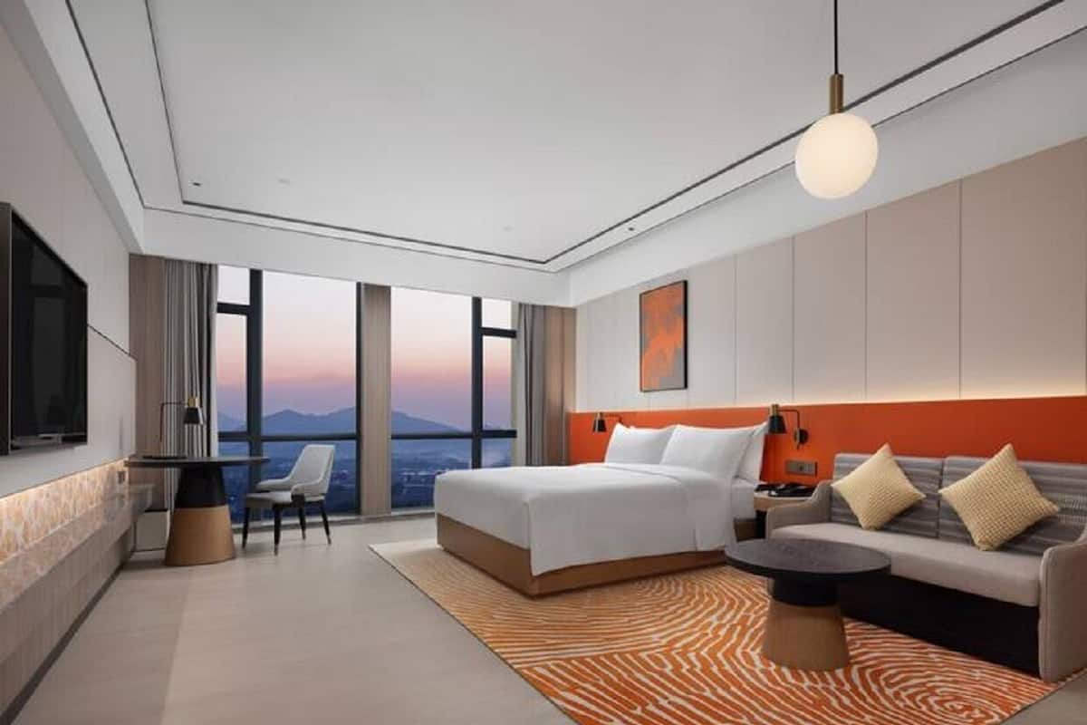 A modern hotel room with a large bed, a sofa, a dining table, and a view of the cityscape.
