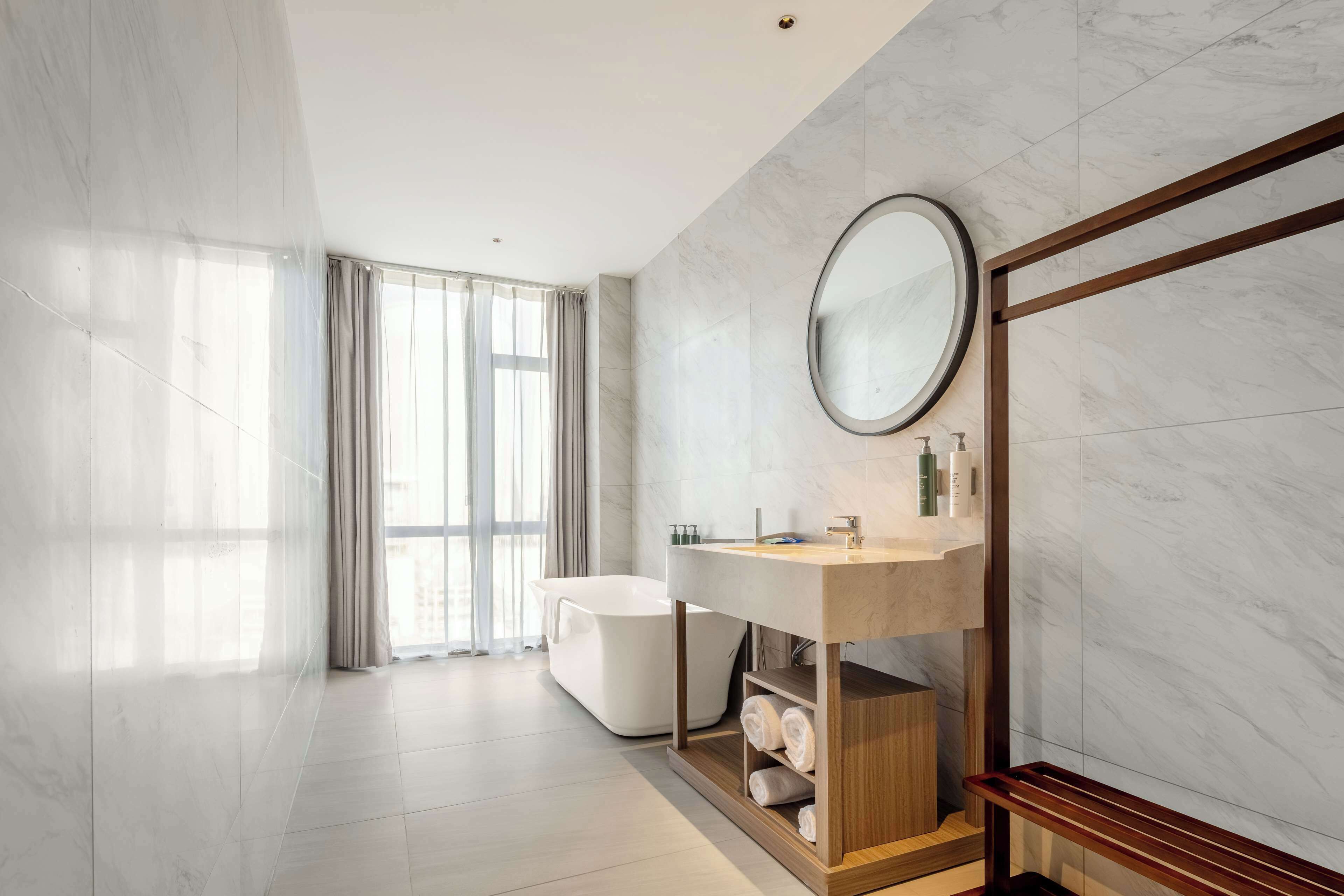 A modern bathroom with a freestanding bathtub, a wooden vanity with a mirror, and a large window with curtains.