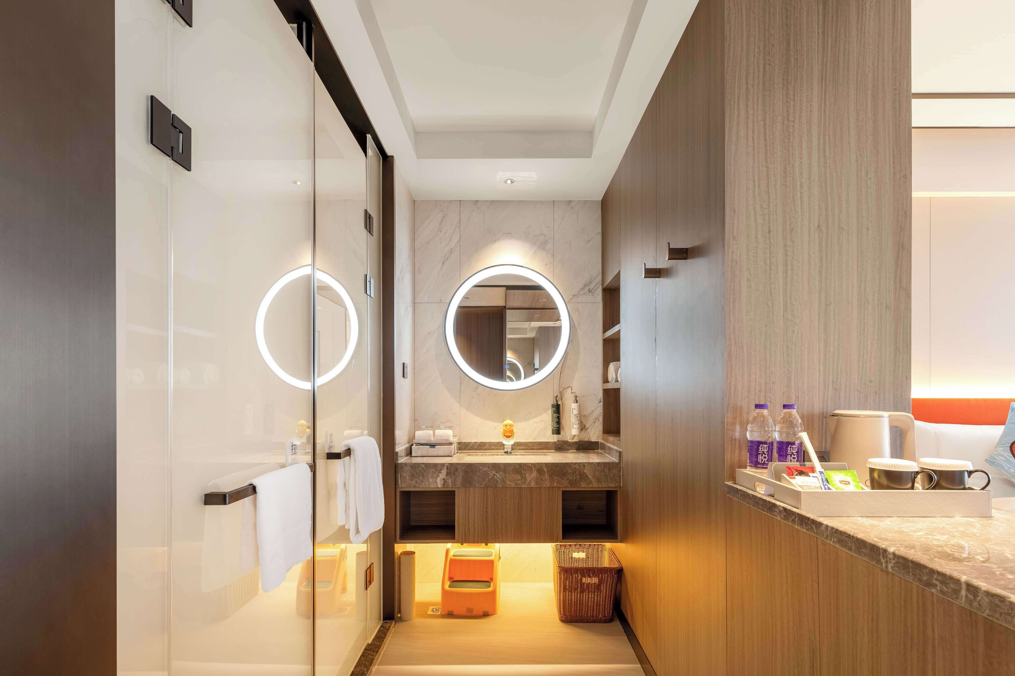 A modern bathroom with a glass partition, a vanity with a circular mirror, and a small seating area with a basket.