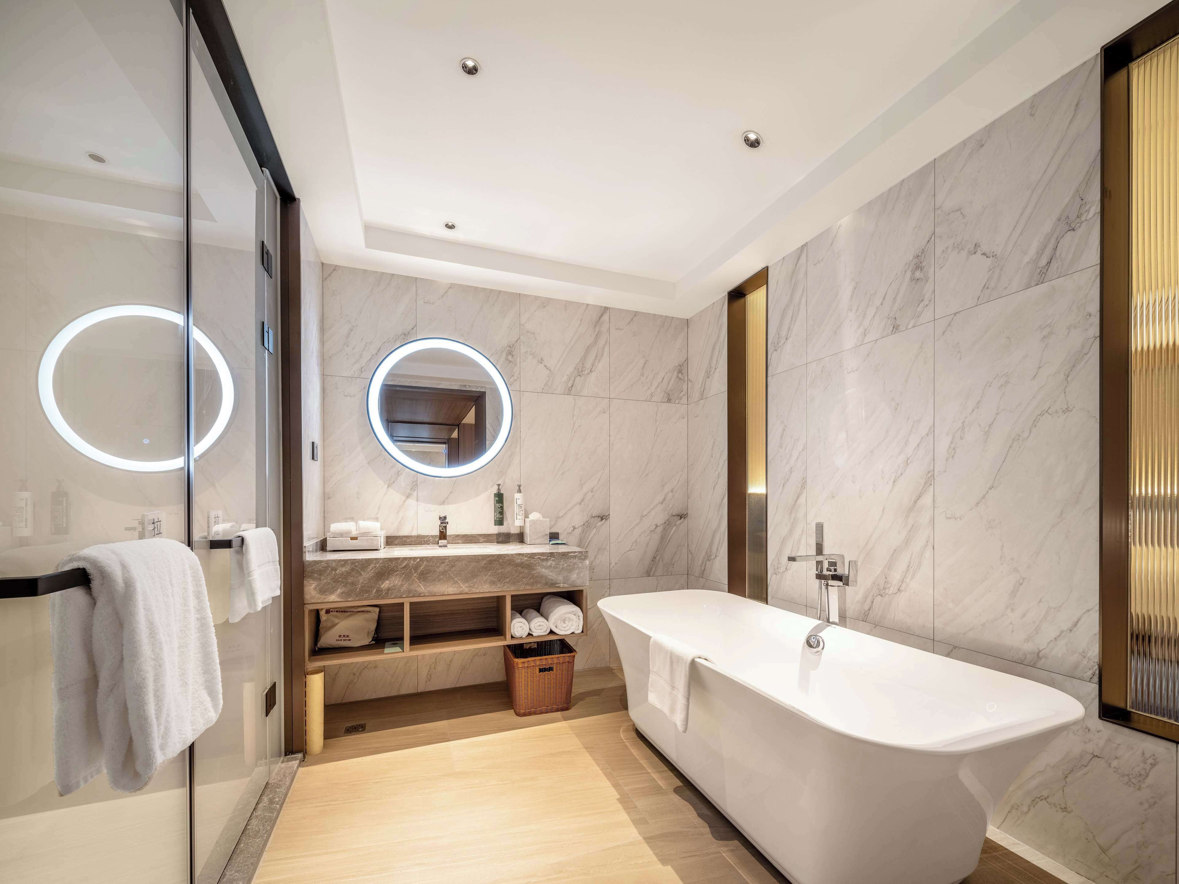 Un baño moderno con una bañera independiente, paredes de mármol, ducha con mampara de vidrio y un tocador con espejo y toallero.