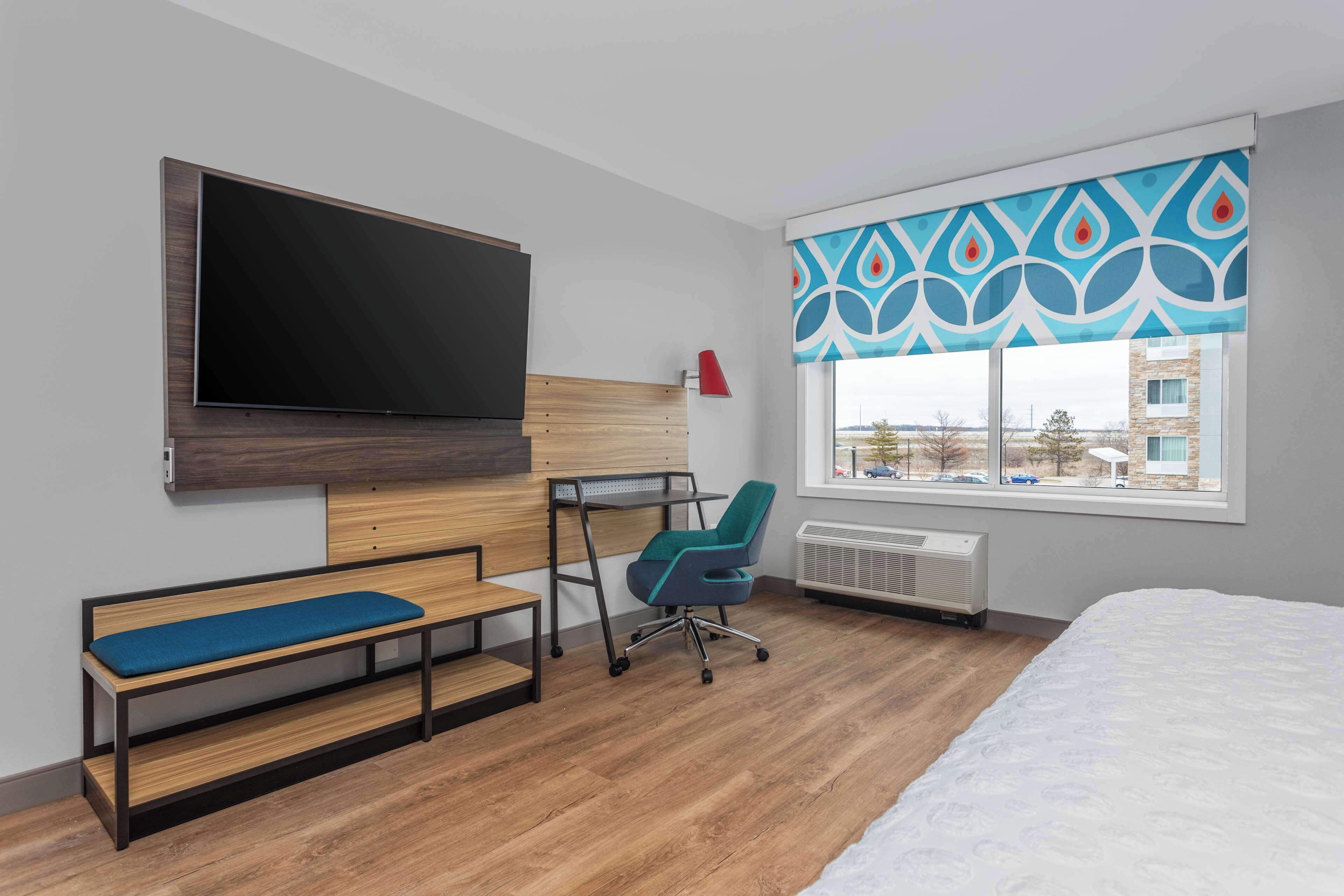 A modern hotel room with a large bed, a desk with a chair, a bench, a TV mounted on the wall, and a window with patterned curtains.
