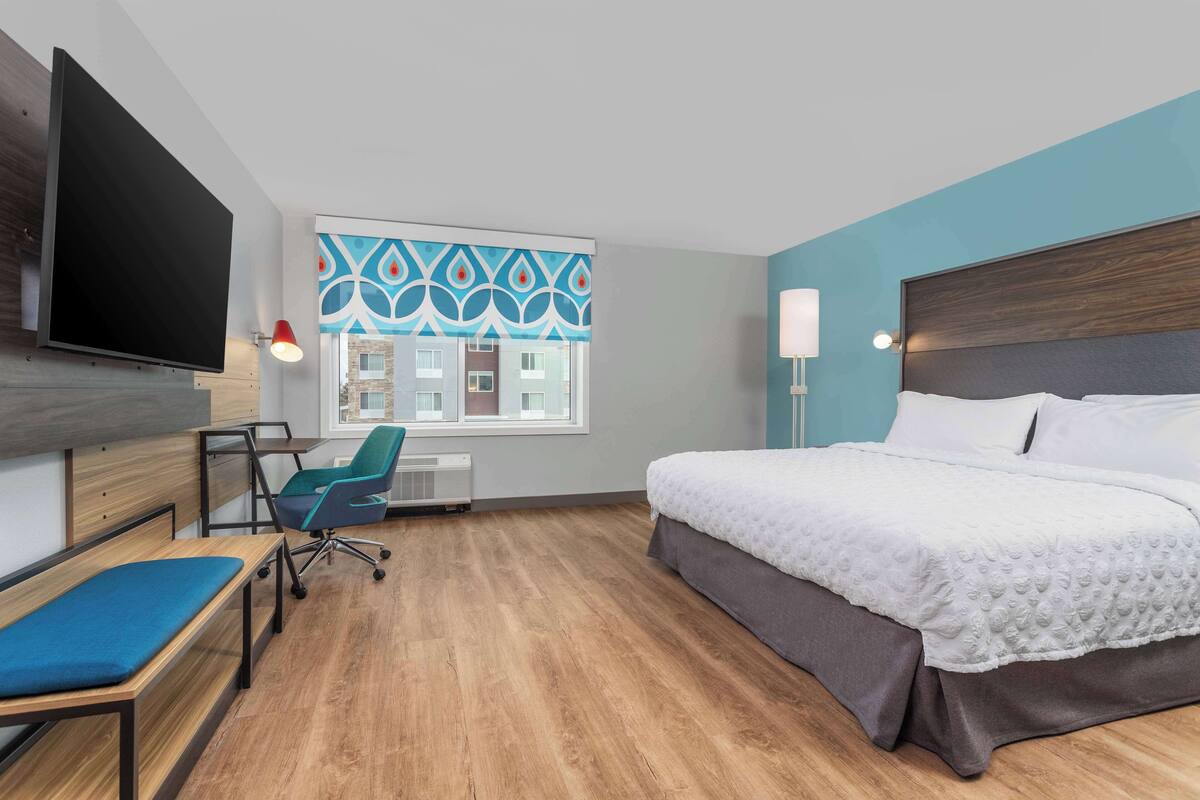 Une chambre d’hôtel moderne dotée d’un grand lit, d’un bureau avec une chaise, d’un téléviseur fixé au mur et d’une fenêtre agrémentée de rideaux à motifs.