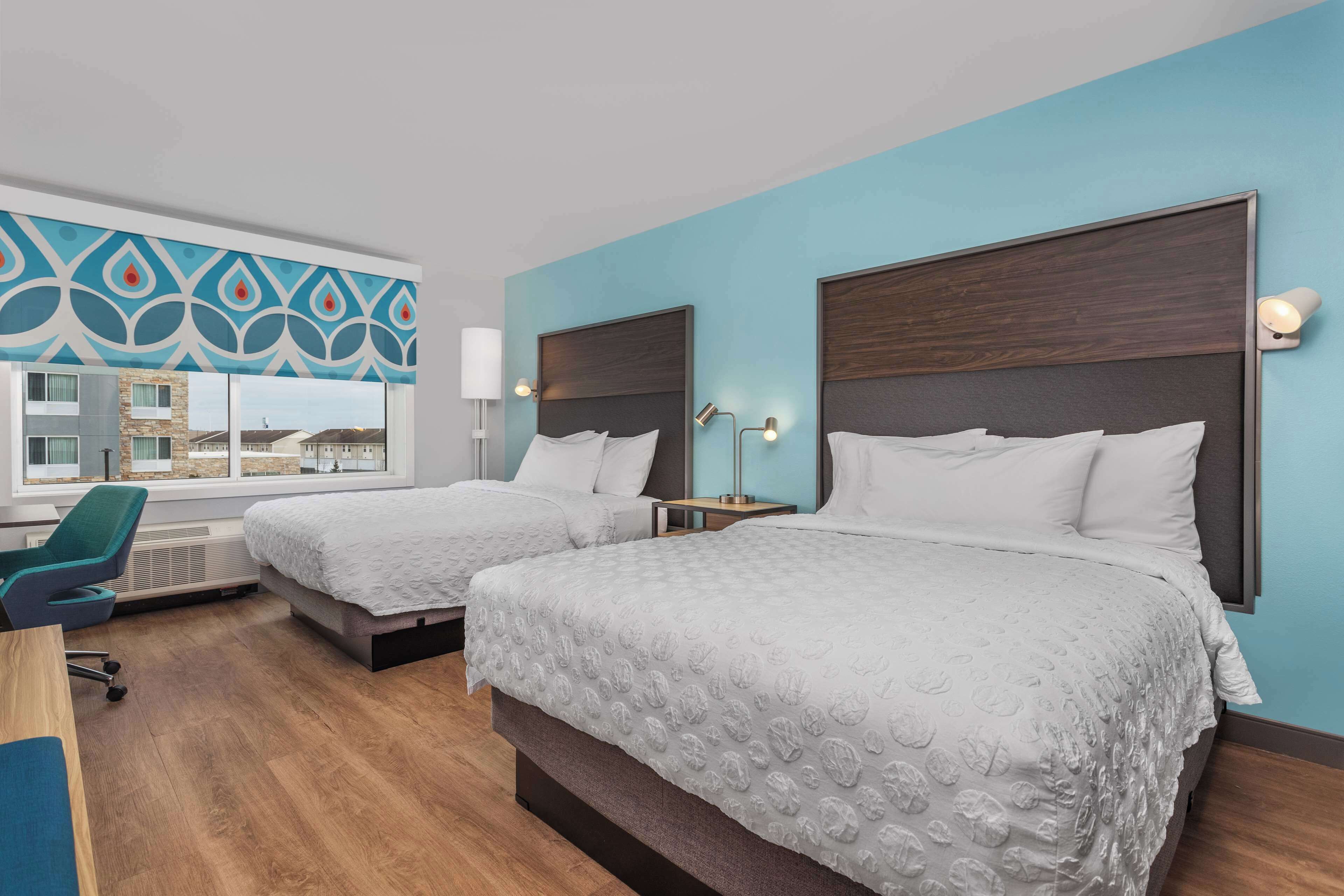 Une chambre d’hôtel avec deux lits, un bureau, une chaise et une grande fenêtre donnant sur des immeubles.
