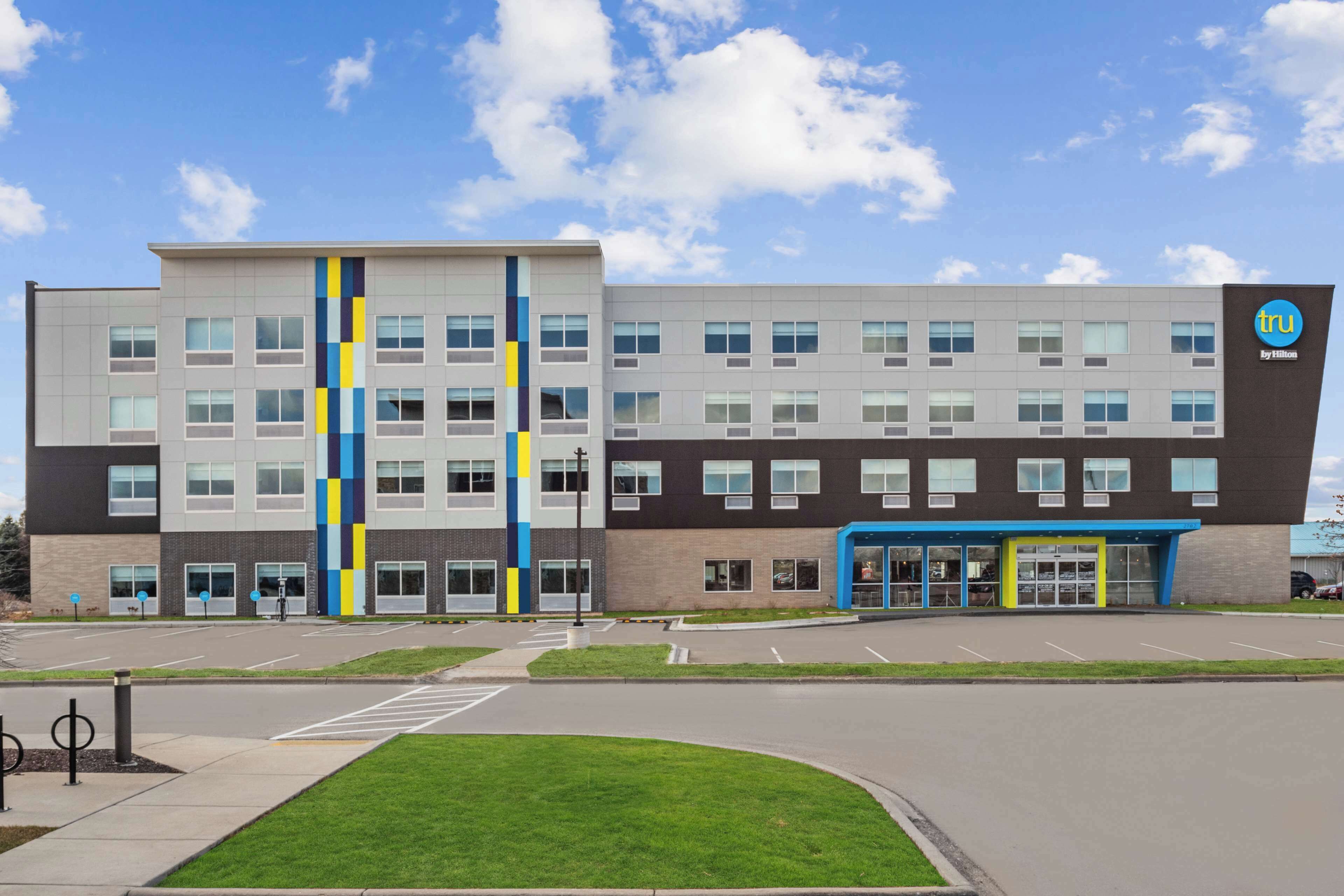A modern building with a mix of grey and brown exterior, a blue entrance, and a logo on the right side.