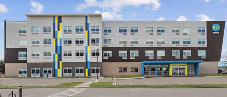 A modern building with a mix of grey and brown exterior, a blue entrance, and a logo on the right side.