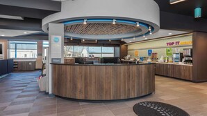 A modern hotel reception area with a wooden reception desk, a 'Top It Breakfast' counter, and a visible recycling bin.