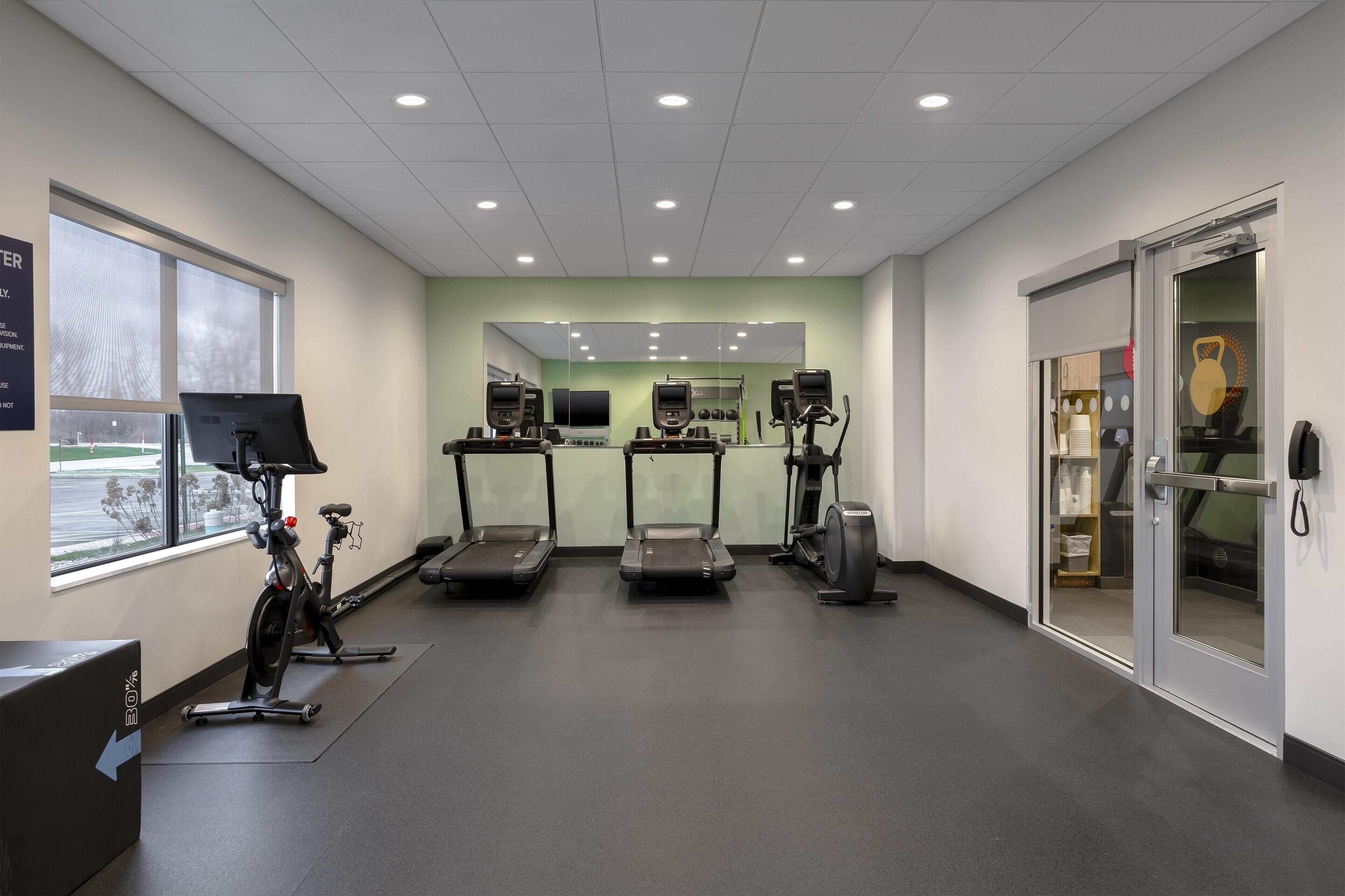 Un gimnasio con cintas de correr, bicicletas estáticas y una máquina de remo. Hay un ventanal grande, un espejo y una puerta que da a un área de almacenamiento.