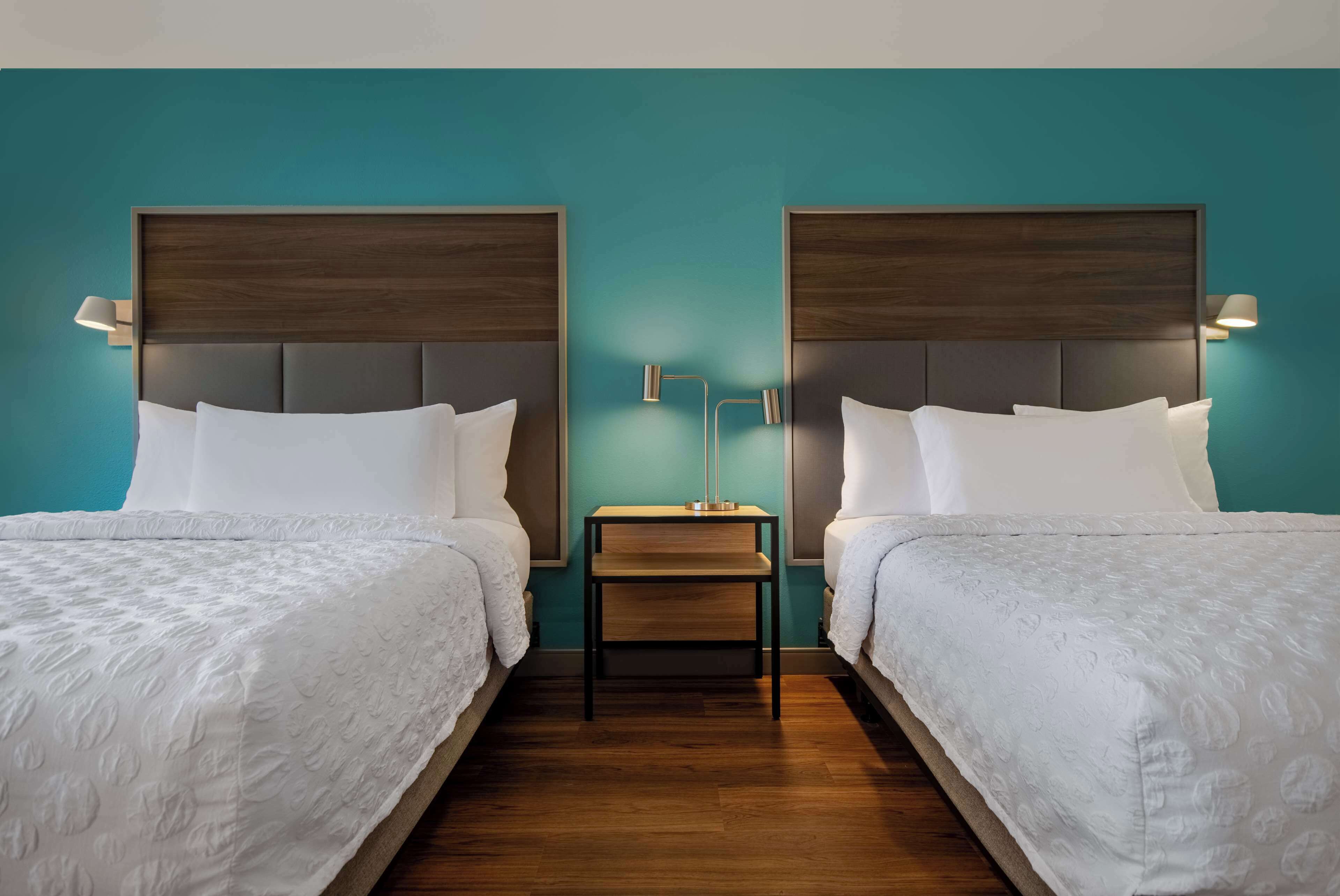Habitación de hotel con dos camas, cabeceros de madera, ropa de cama blanca y una mesita de noche con lámpara.
