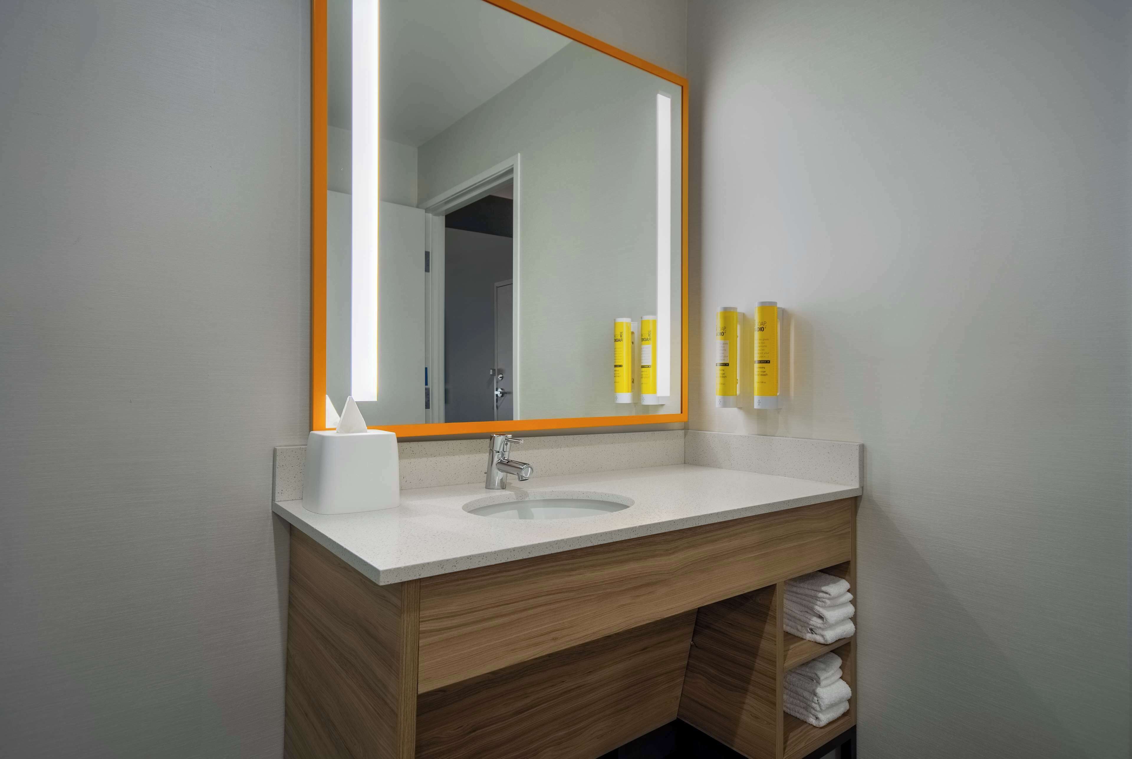 Ein modernes Badezimmer mit einem weißen Waschbecken, einer Holzkommode und einem großen Spiegel mit orangefarbenem Rahmen.