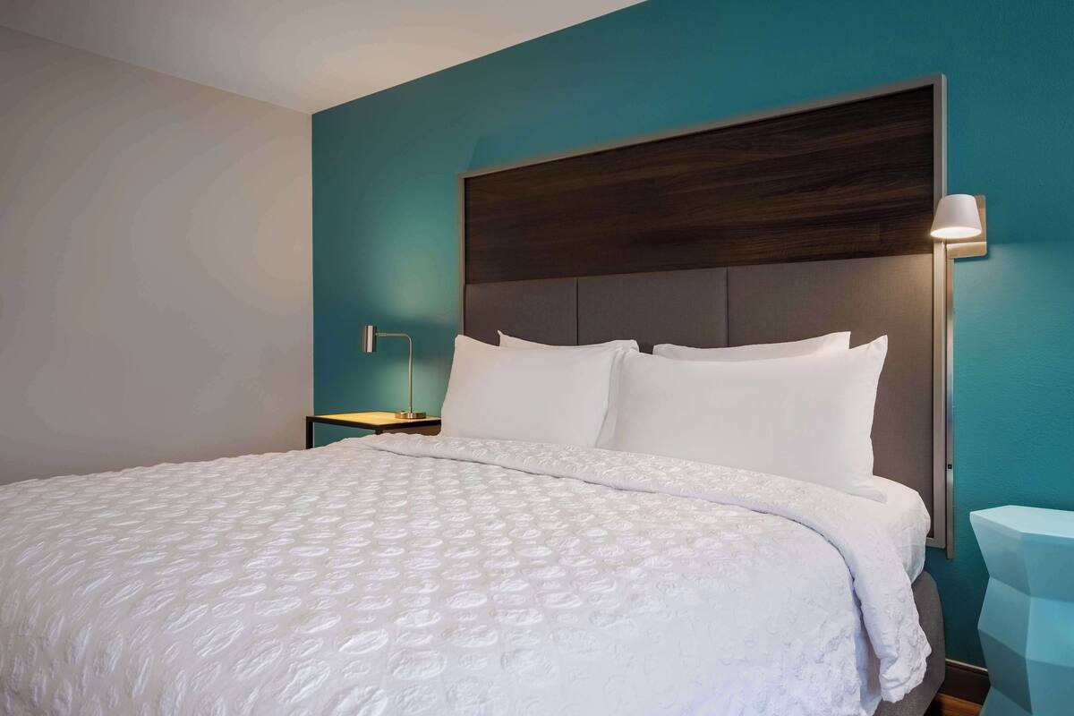 Un lit bien fait, avec un couvre-lit blanc et des oreillers, une tête de lit en bois foncé et une table de chevet avec une lampe.