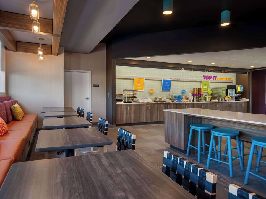 A modern restaurant with wooden tables and blue bar stools, a counter with kitchen equipment, and a seating area with a sofa and cushions.