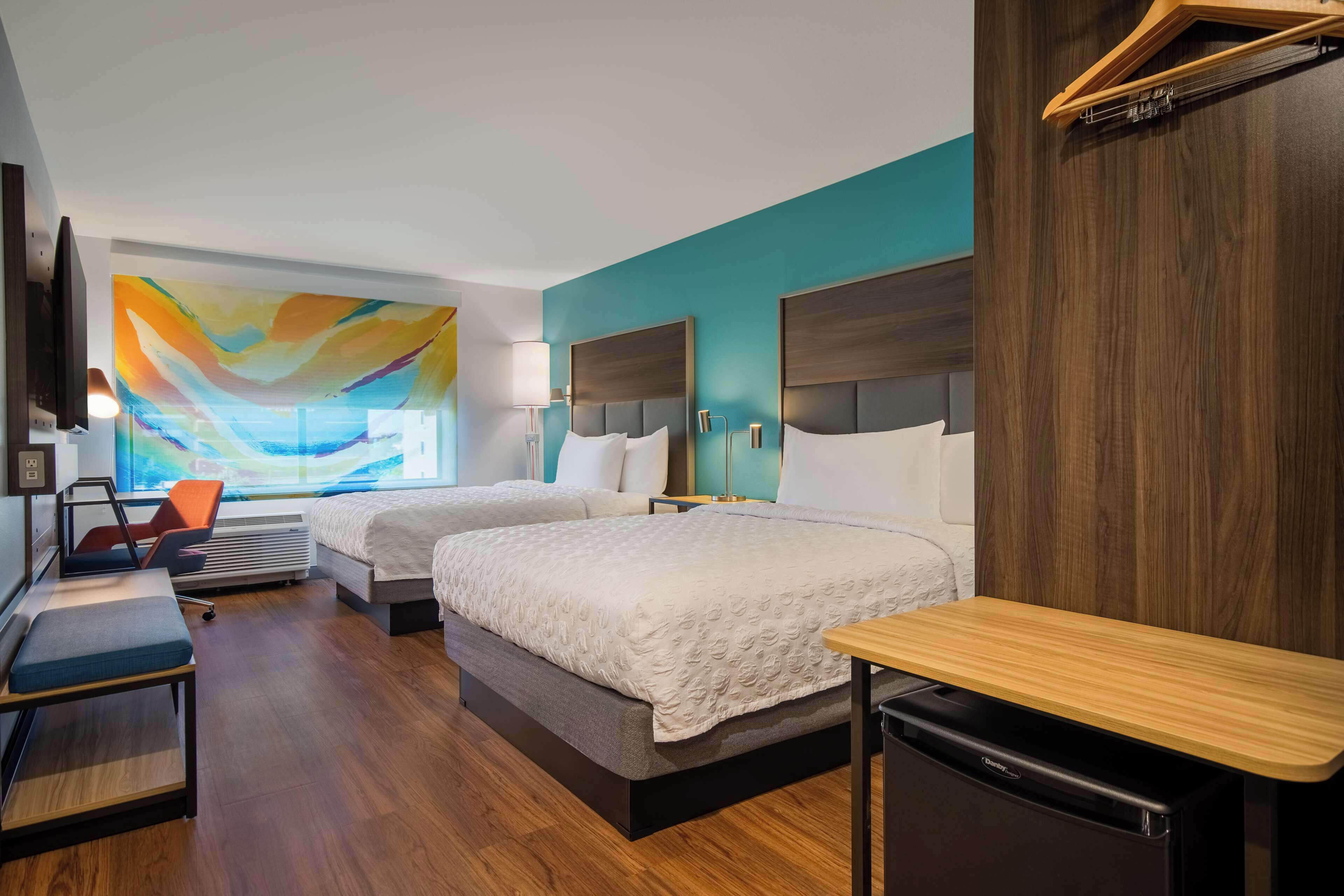 Habitación de hotel con dos camas, un escritorio, una silla, un televisor y un cuadro abstracto grande en la pared.