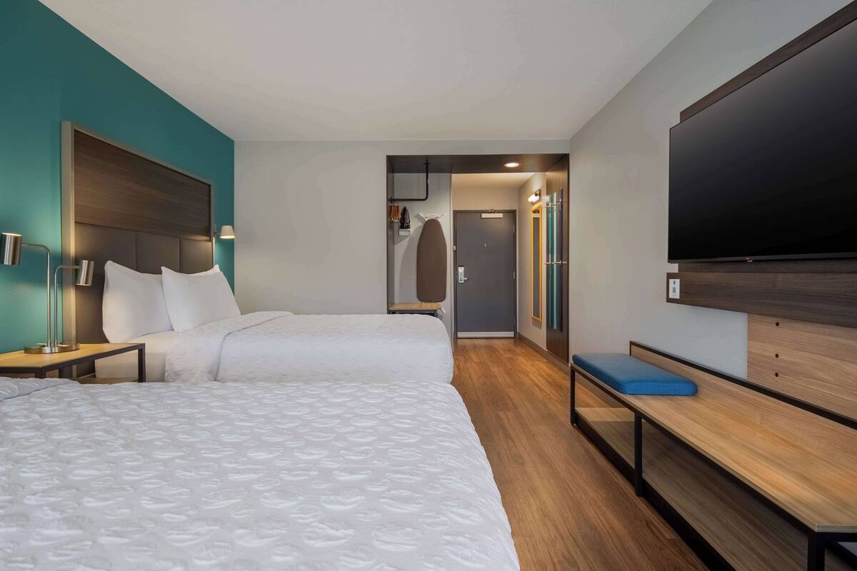 Une chambre d’hôtel avec deux lits, un banc, une télévision et une porte donnant sur une salle de bain.
