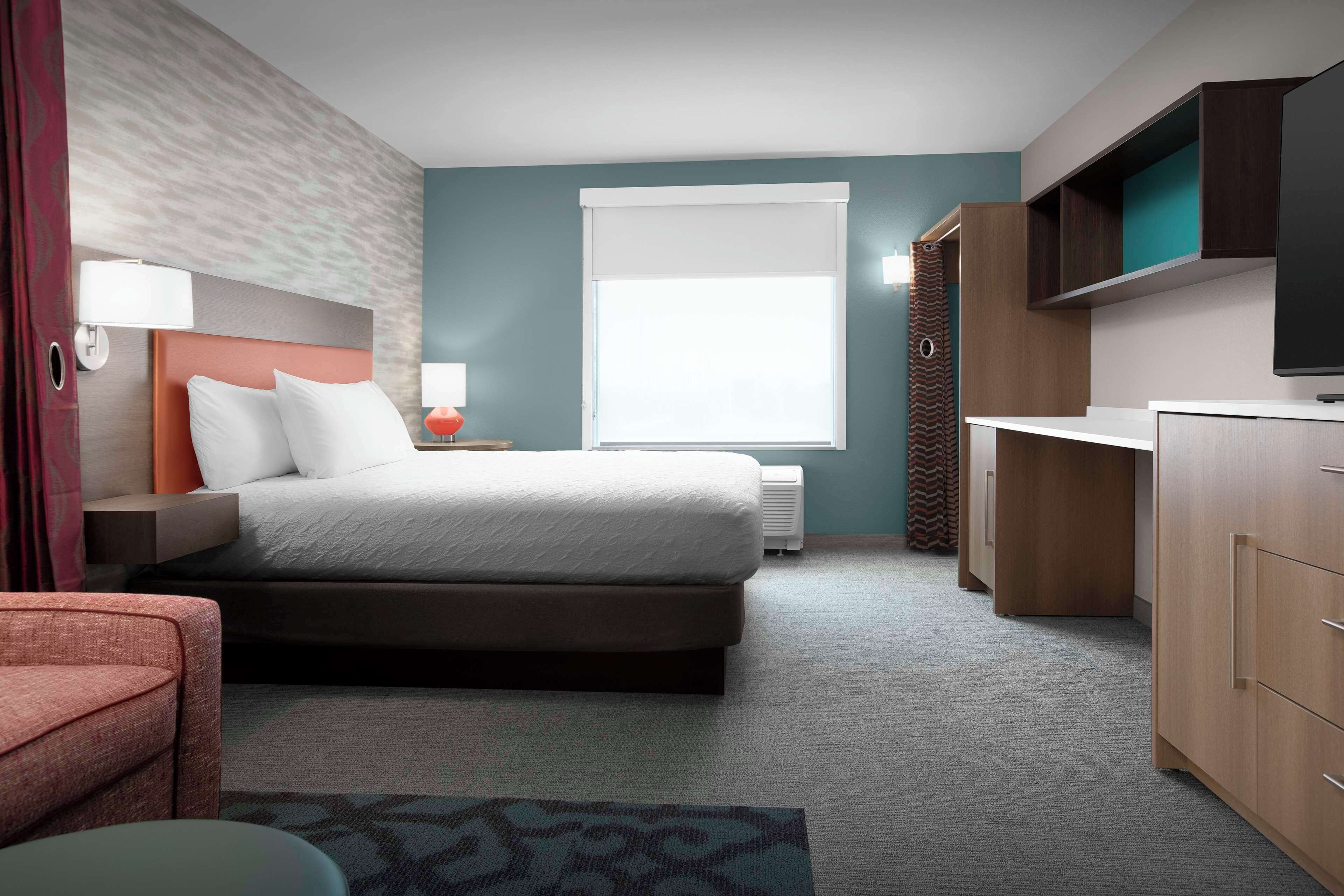 Quarto de hotel moderno com cama, mesa de cabeceira, abajur e televisão fixada na parede.