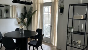 Un espace de salle à manger moderne avec une table ronde, des chaises noires et un lustre noir. On y trouve une fenêtre avec des rideaux, une plante et une étagère avec des bougies et des livres.