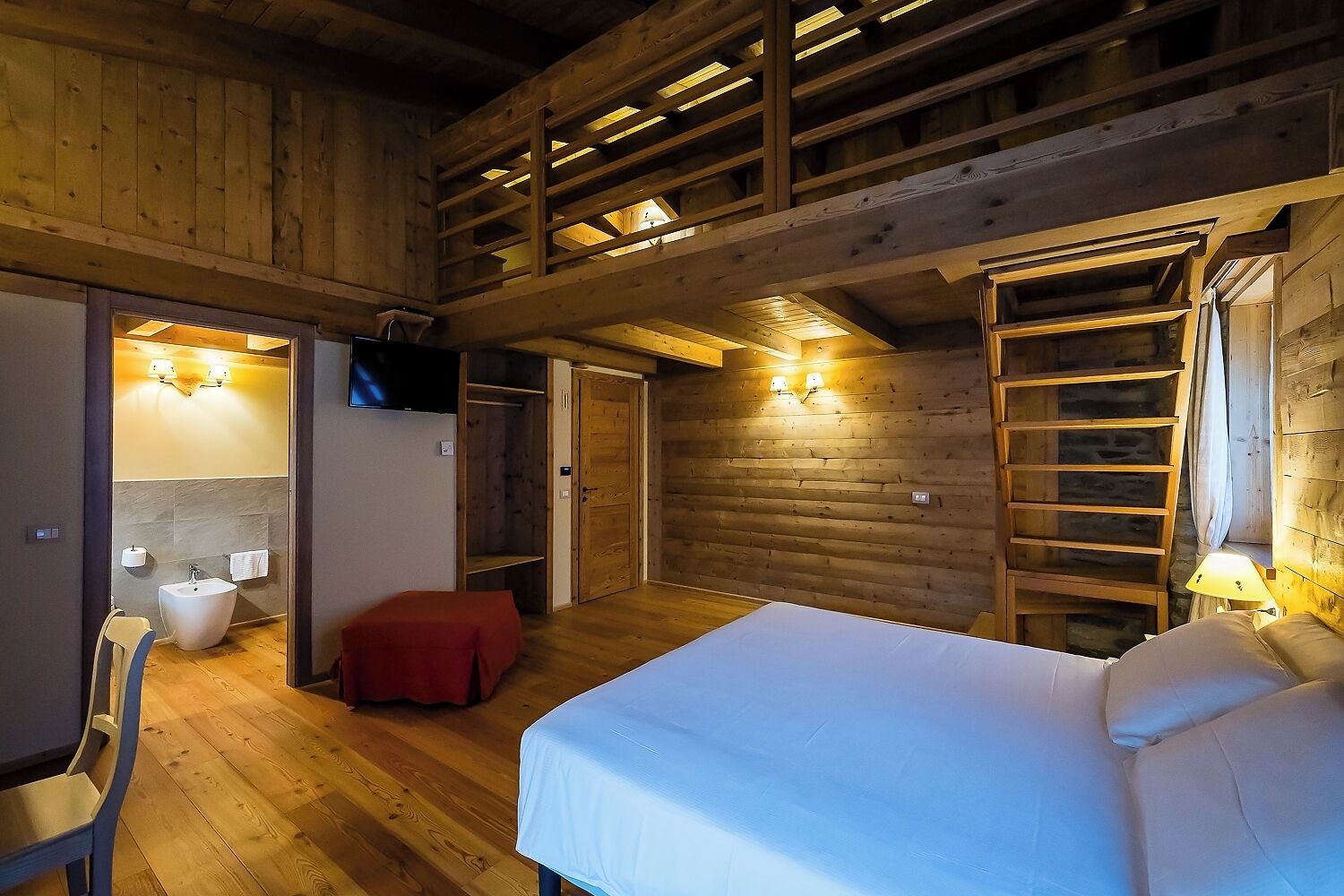 A wooden cabin interior with a bed, a red ottoman, a bathroom with a sink and mirror, and a TV mounted on the wall.