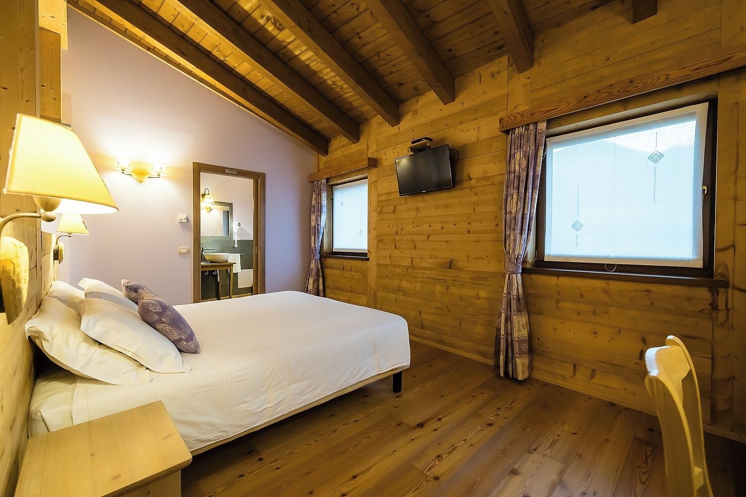 A wooden cabin bedroom with a bed, a nightstand, a lamp, a mirror, a sink, and a window with curtains.