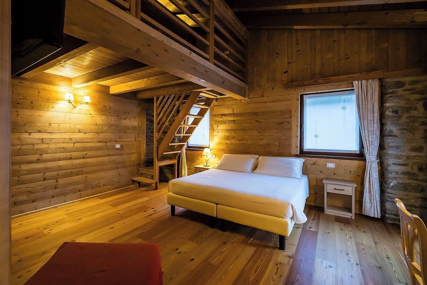 A cozy wooden cabin room with a bed, a small table, a window with curtains, and a staircase.