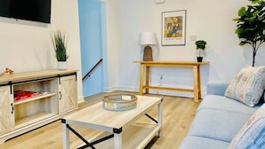 A living room with a sofa, a coffee table, a TV, a lamp, a painting, and a plant. - Updated Home Walkable to Oceanfront! (Virginia Beach)