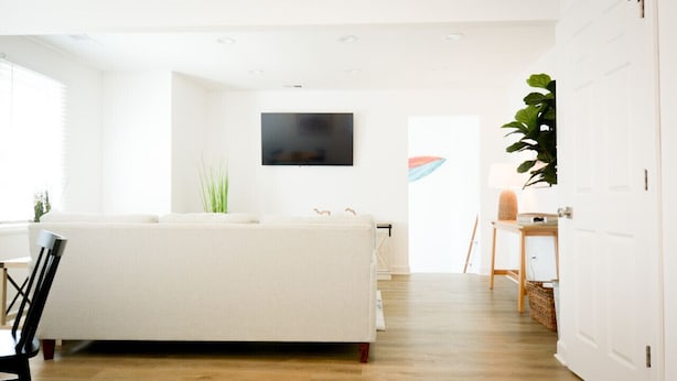 A modern living room with a white sofa, a flat-screen TV mounted on the wall, and a wooden floor. - Updated Home Walkable to Oceanfront! (Virginia Beach)