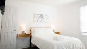 A neatly arranged bedroom with a bed, bedside lamps, a wall-mounted artwork, and a window with blinds. - Updated Home Walkable to Oceanfront! (Virginia Beach)