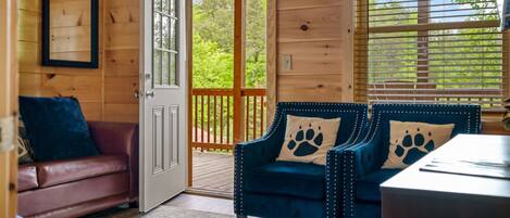 A cozy cabin interior with a wooden door, a blue armchair with paw prints, a brown leather sofa, and a window with blinds.