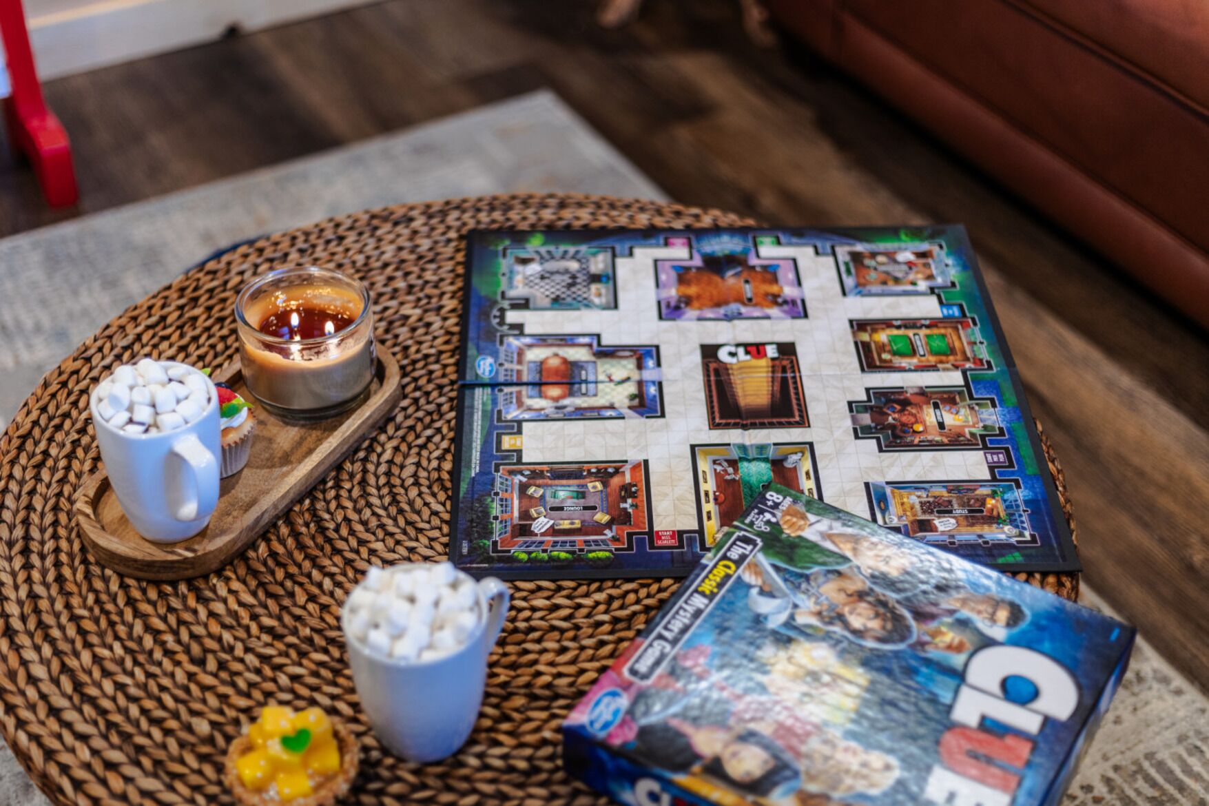 A board game with a map layout, a mug with marshmallows, a candle, and a snack bowl on a woven mat.