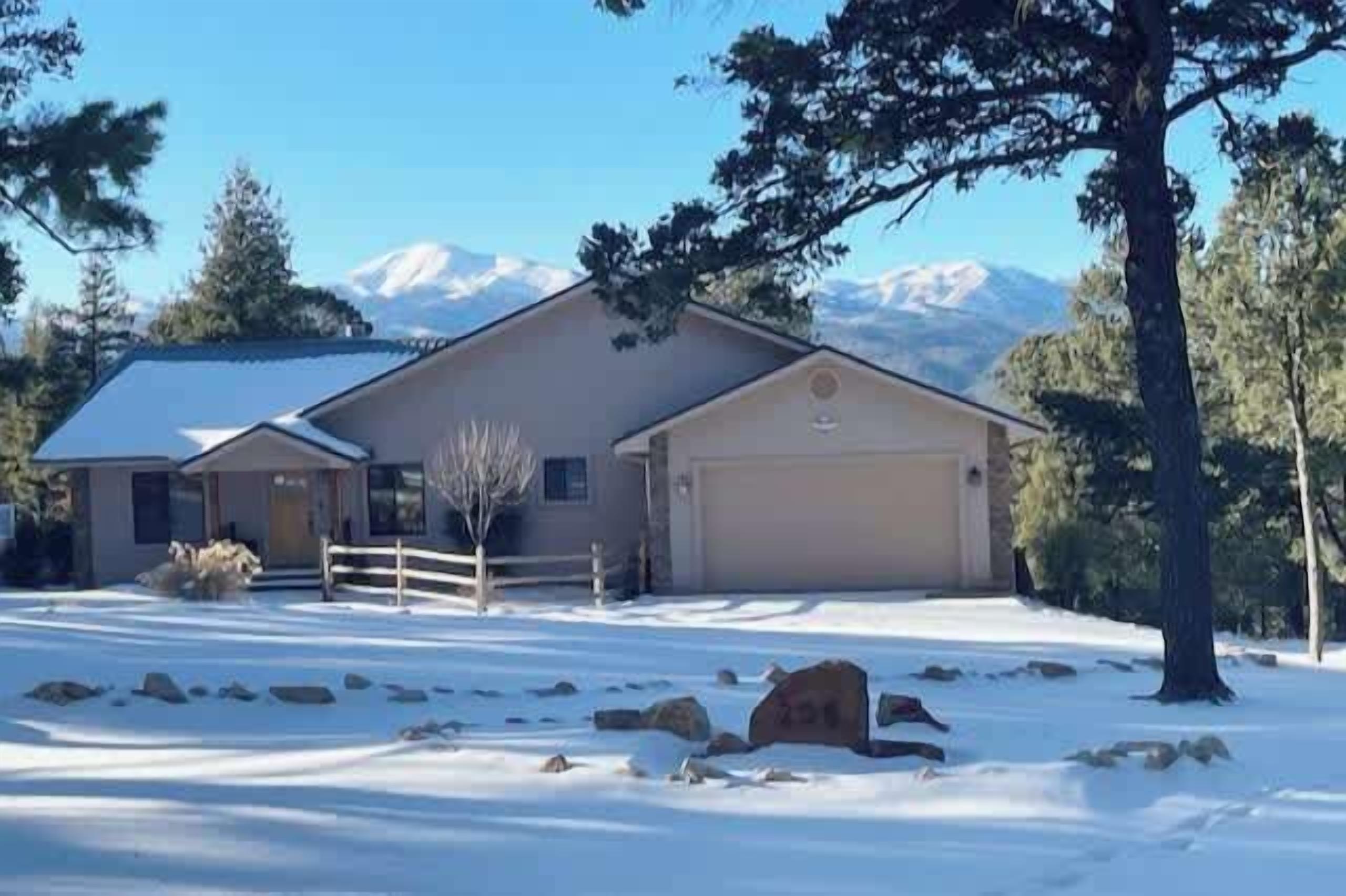 A two-story house with a garage, surrounded by snow-covered ground and trees, with mountains in the background.