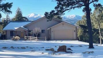 A two-story house with a garage, surrounded by snow-covered ground and trees, with mountains in the background.