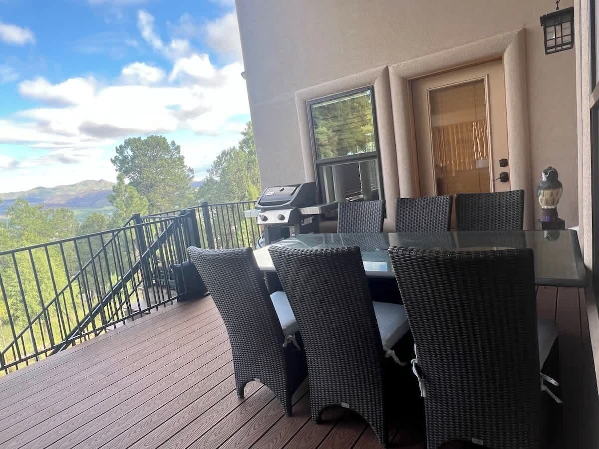 A deck with a glass table and wicker chairs, overlooking a scenic landscape.