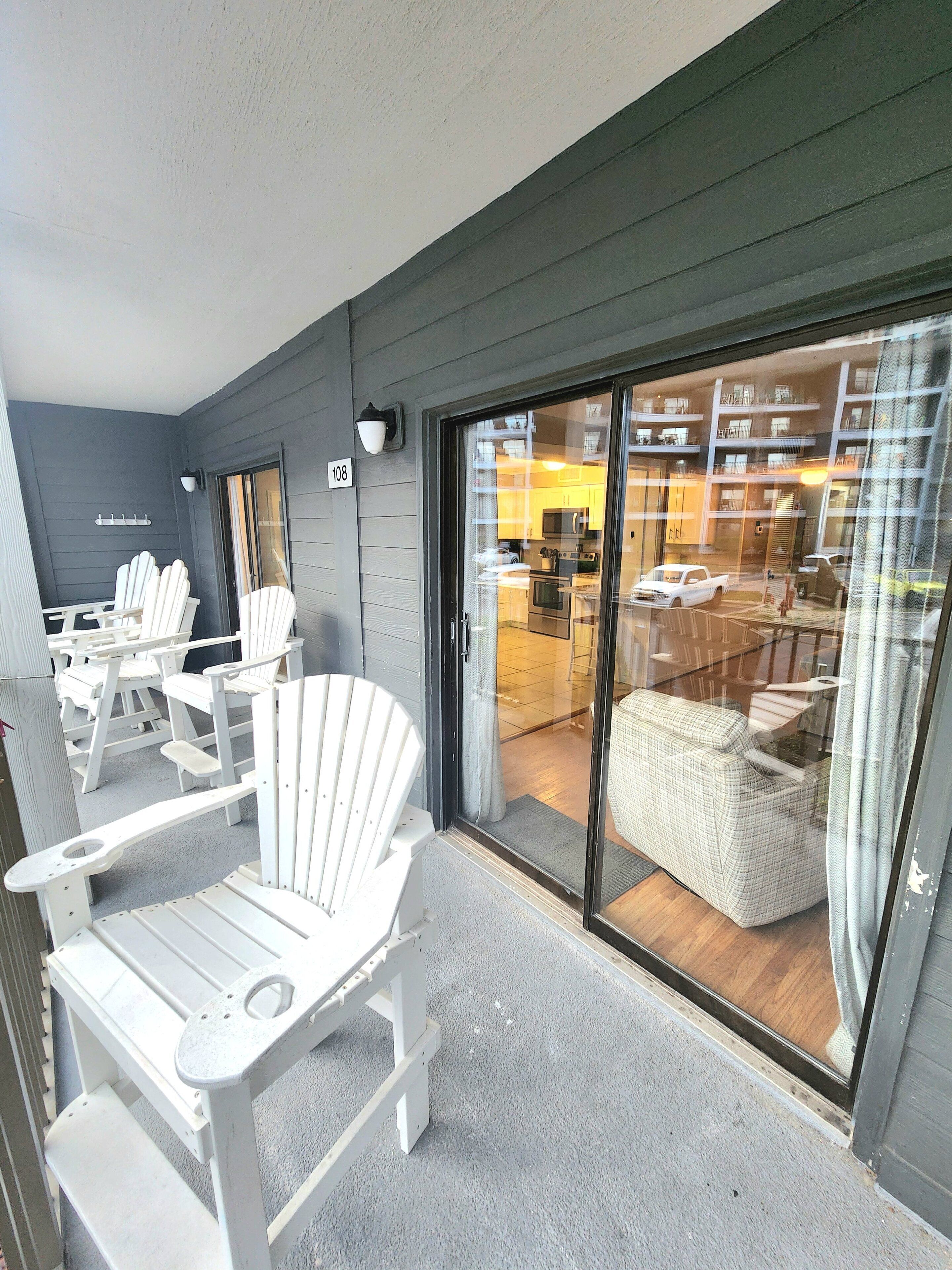 A balcony with white Adirondack chairs and a sliding glass door leading to a modern interior with a kitchen and seating area.