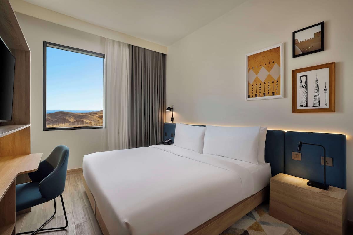 Ein Hotelzimmer mit einem großen Bett, einem Schreibtisch, einem Sessel, einem Fernseher und einem Fenster mit Blick auf die Hügel.