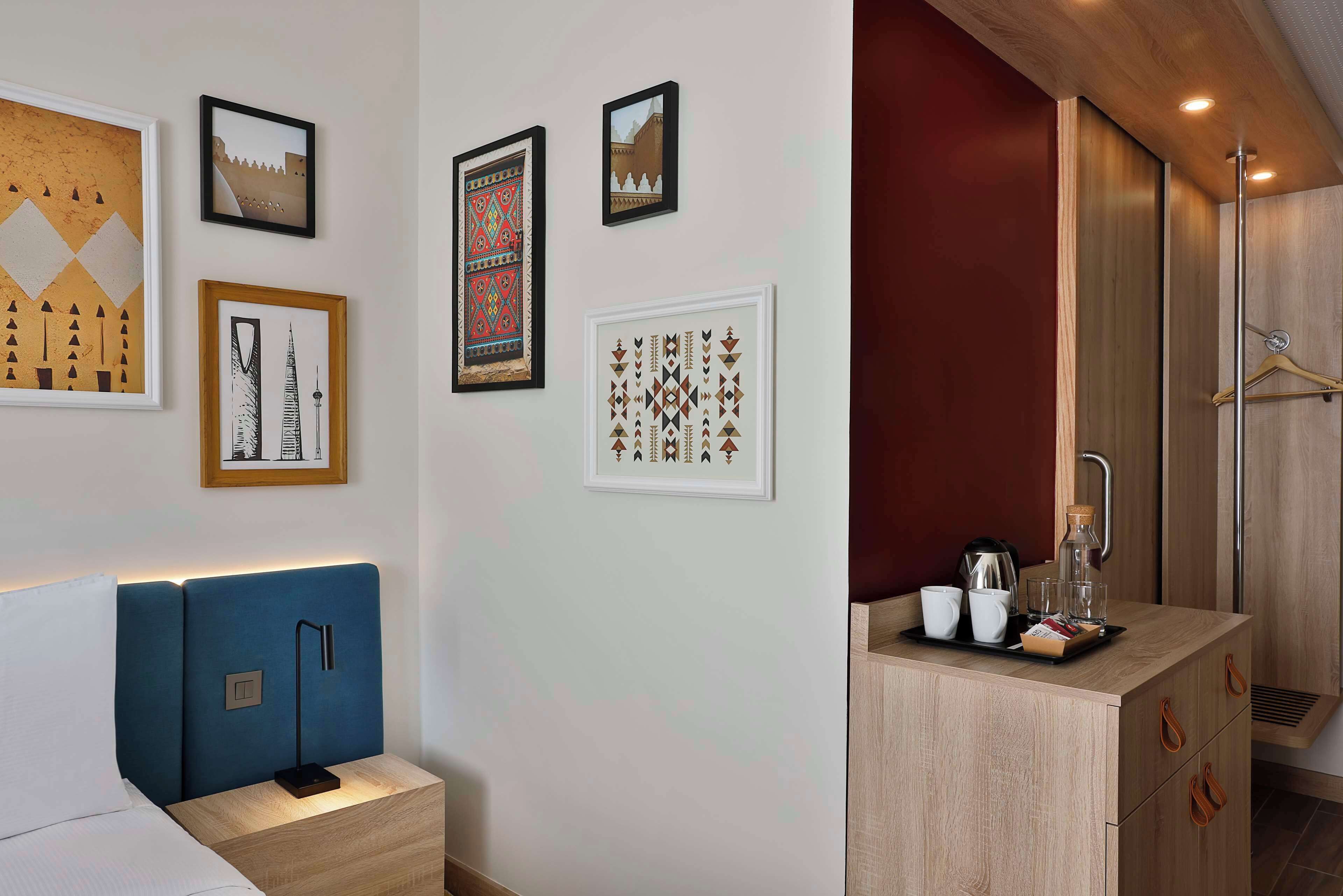 Une chambre d’hôtel moderne équipée d’un lit, d’une table de chevet, d’œuvres d’art murales et d’une petite kitchenette avec un plateau contenant divers articles.