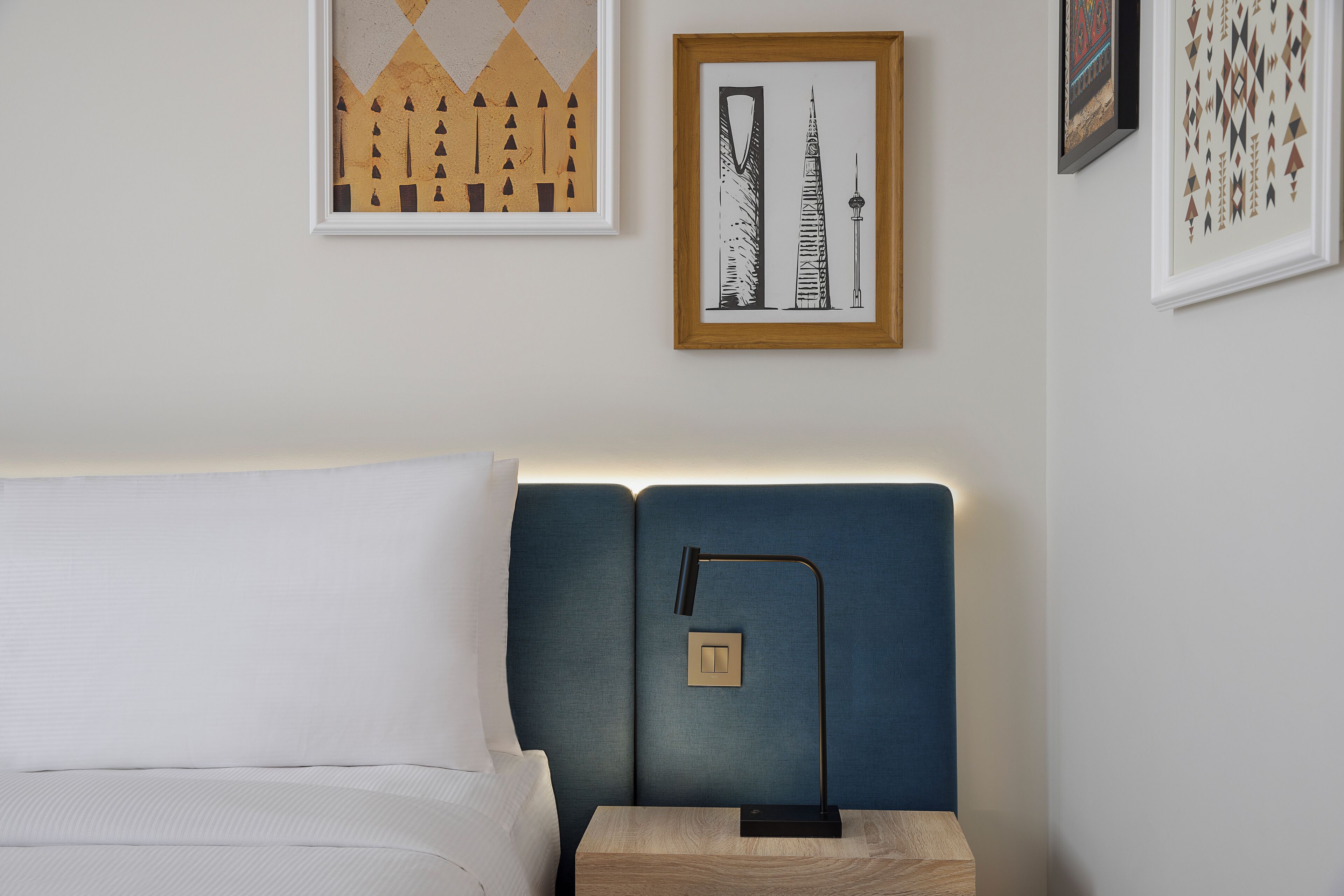 A neatly made bed with a white comforter and pillows, a blue upholstered headboard, a bedside table with a lamp, and framed artwork on the wall.