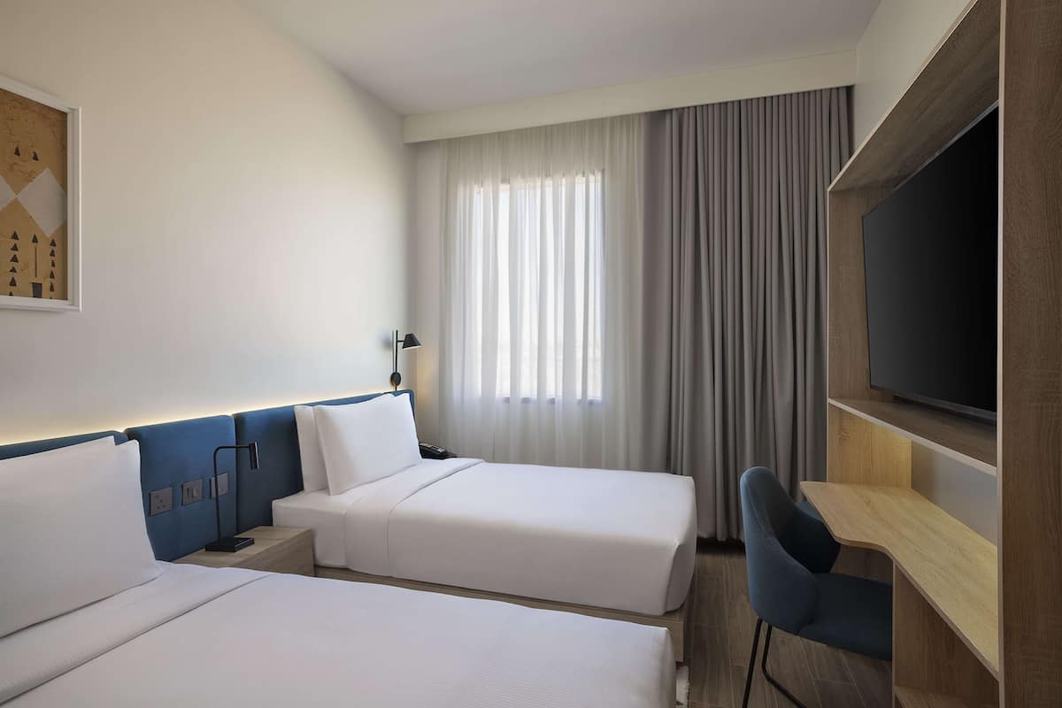 Ein Hotelzimmer mit zwei Betten, einem Schreibtisch, einem Fernseher und einem Fenster mit Vorhängen.