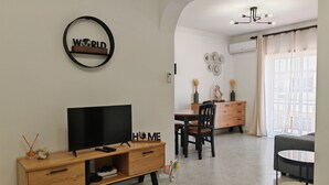 A modern living room with a TV, wooden furniture, a dining area, and a large window with curtains. - Cristina Apartment - Near the Beach (Armação de Pêra)