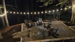 Un espacio al aire libre con asientos tipo Adirondack y un hogar para fogata, rodeado de árboles y luces colgantes.