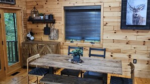 Interior de una cabaña de madera con una mesa, bancos, una ventana con persianas, un cuadro de un ciervo colgado y una puerta con la leyenda 'ARRO CREEK'.