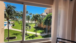 A view from a balcony overlooking a pool area with palm trees and a building with a thatched roof. - Best Location 3BR Beachfront Condo with Ocean View (Puerto Aventuras)