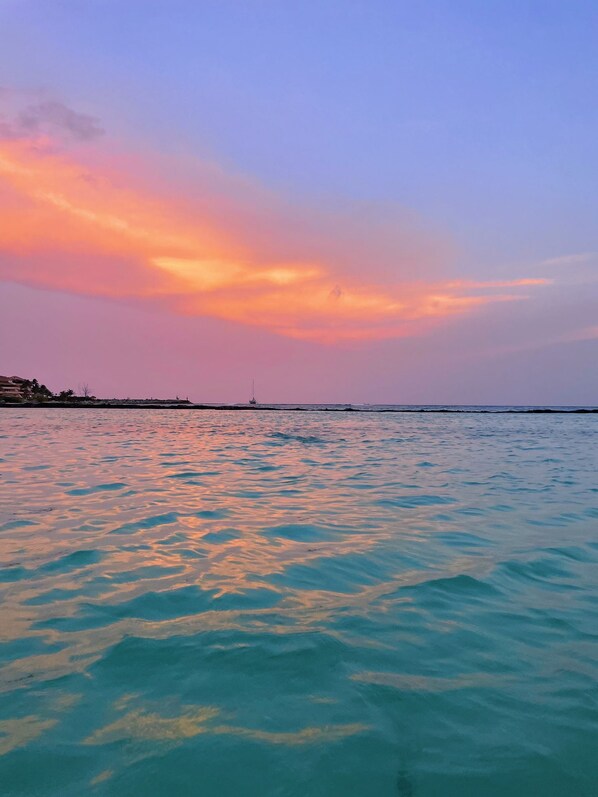 Vista costeira com um pôr do sol sobre o oceano, uma linha de costa distante e um pequeno barco.