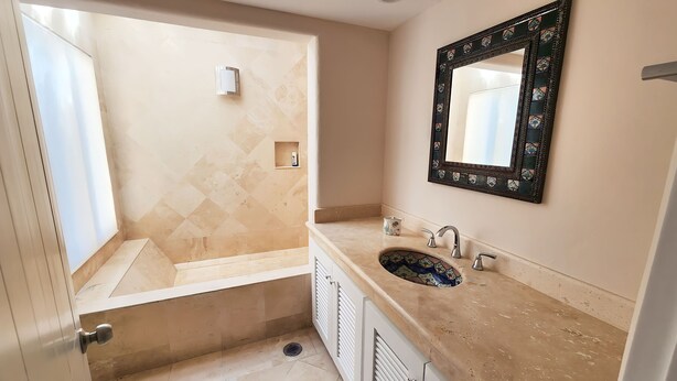 A bathroom with a beige tiled wall, a sink with a decorative mirror, and a shower area. - Best Location 3BR Beachfront Condo with Ocean View (Puerto Aventuras)