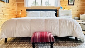 A wooden cabin bedroom with a bed, a nightstand, a bench, and a window with curtains. - Sleeping Lion Casita in Fort Davis, Texas. Explore The Davis Mountains and area. (Fort Davis)
