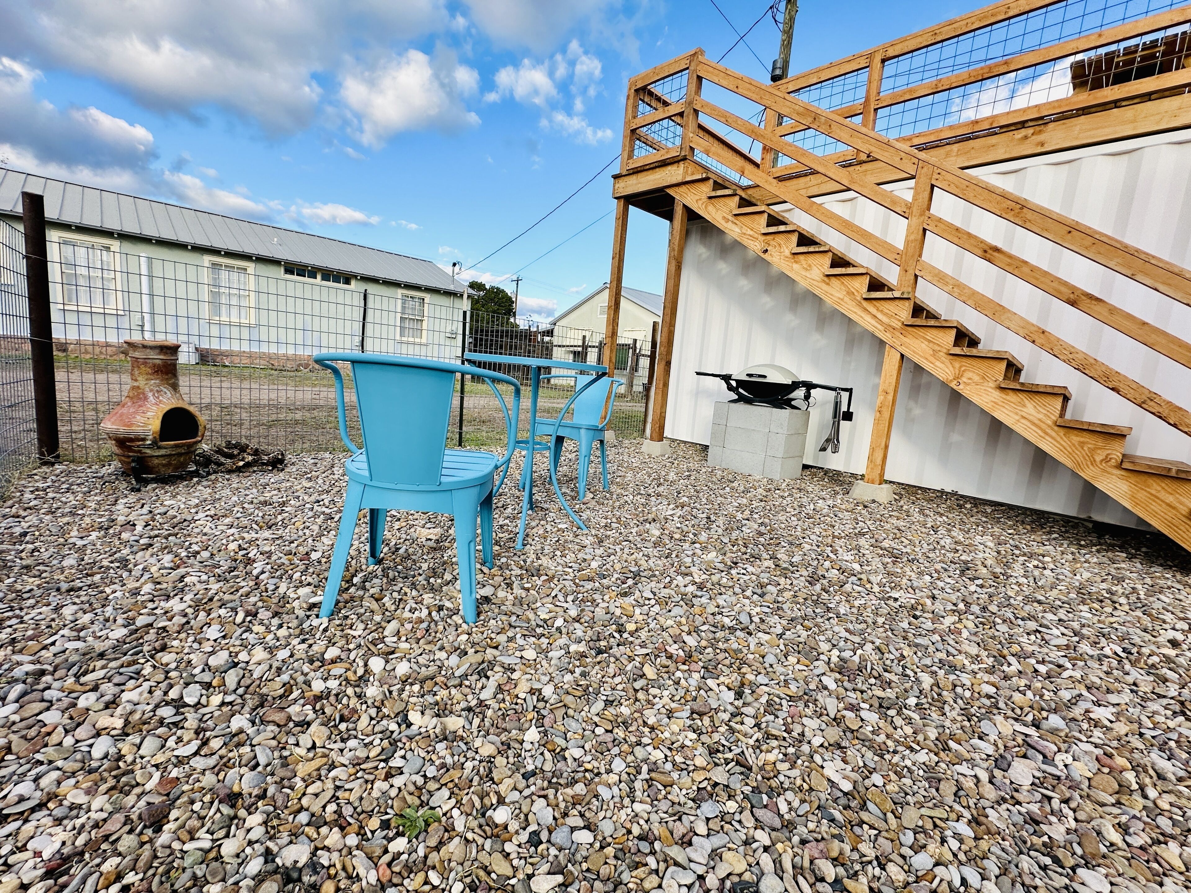 A patio area with a wooden staircase, a grill, and blue chairs.
