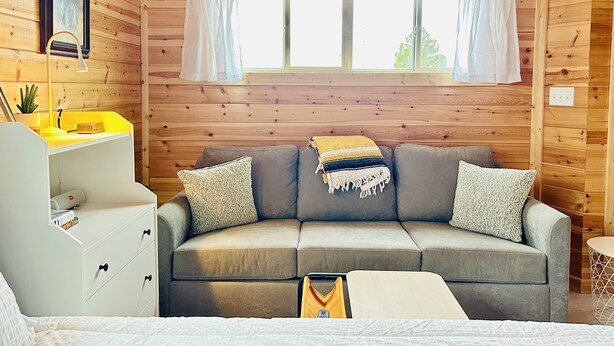 A cozy room with a grey sofa, a small white desk, and a window with white curtains. - Sleeping Lion Casita in Fort Davis, Texas. Explore The Davis Mountains and area. (Fort Davis)