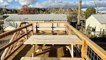 Une terrasse en bois avec une table de pique-nique et une clôture, donnant sur un quartier résidentiel avec des maisons et une colline rocheuse en arrière-plan.